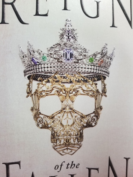 A delicate golden mask in the shape of a skull with ornate bird and bone details, wearing an elaborate jeweled silver crown, pictured on the cover of Sarah Glenn Marsh's novel.